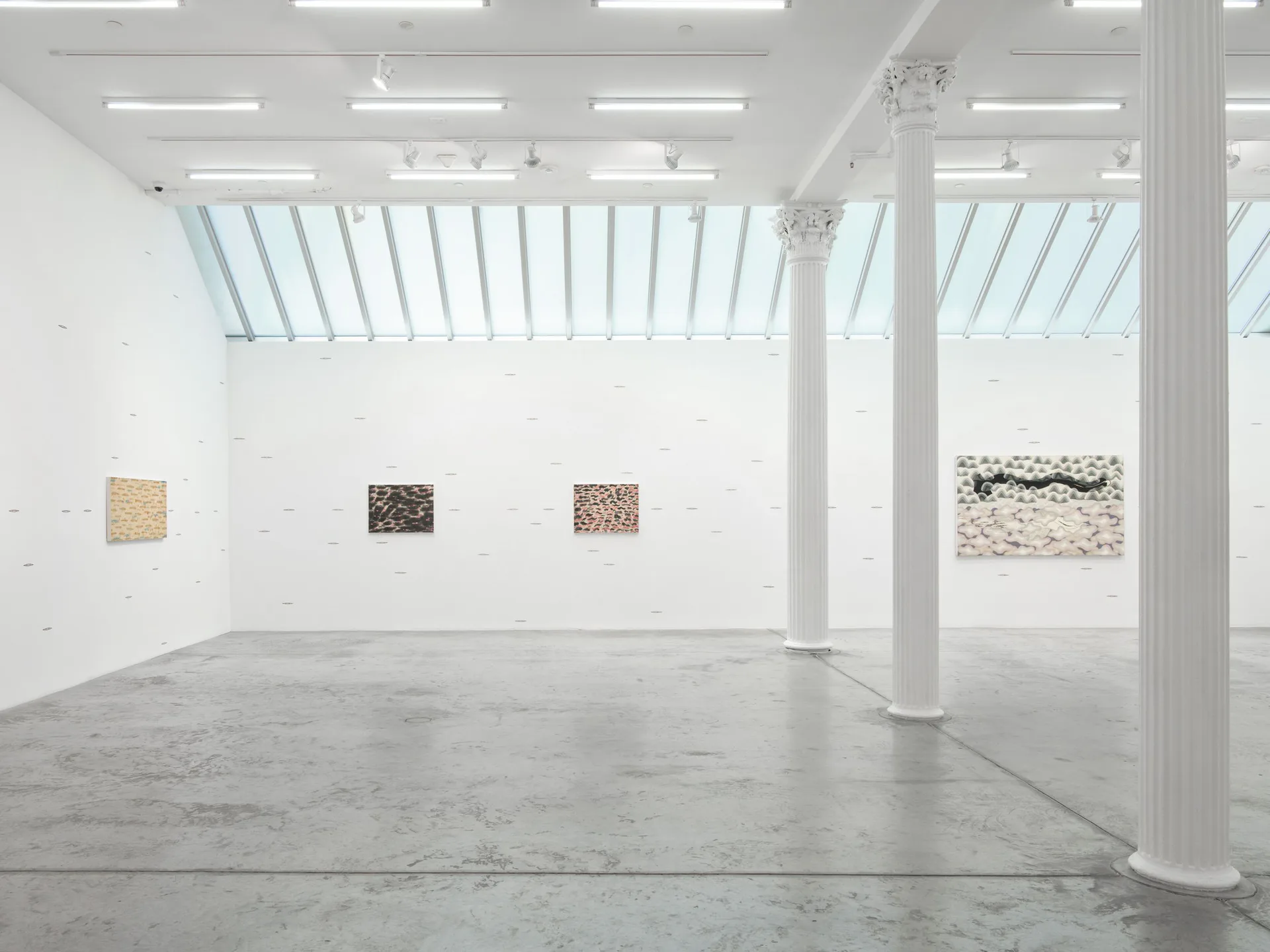 Minimalist gallery interior with polished concrete floor, white walls, skylights, and four abstract textile or mixed-media artworks displayed on walls.