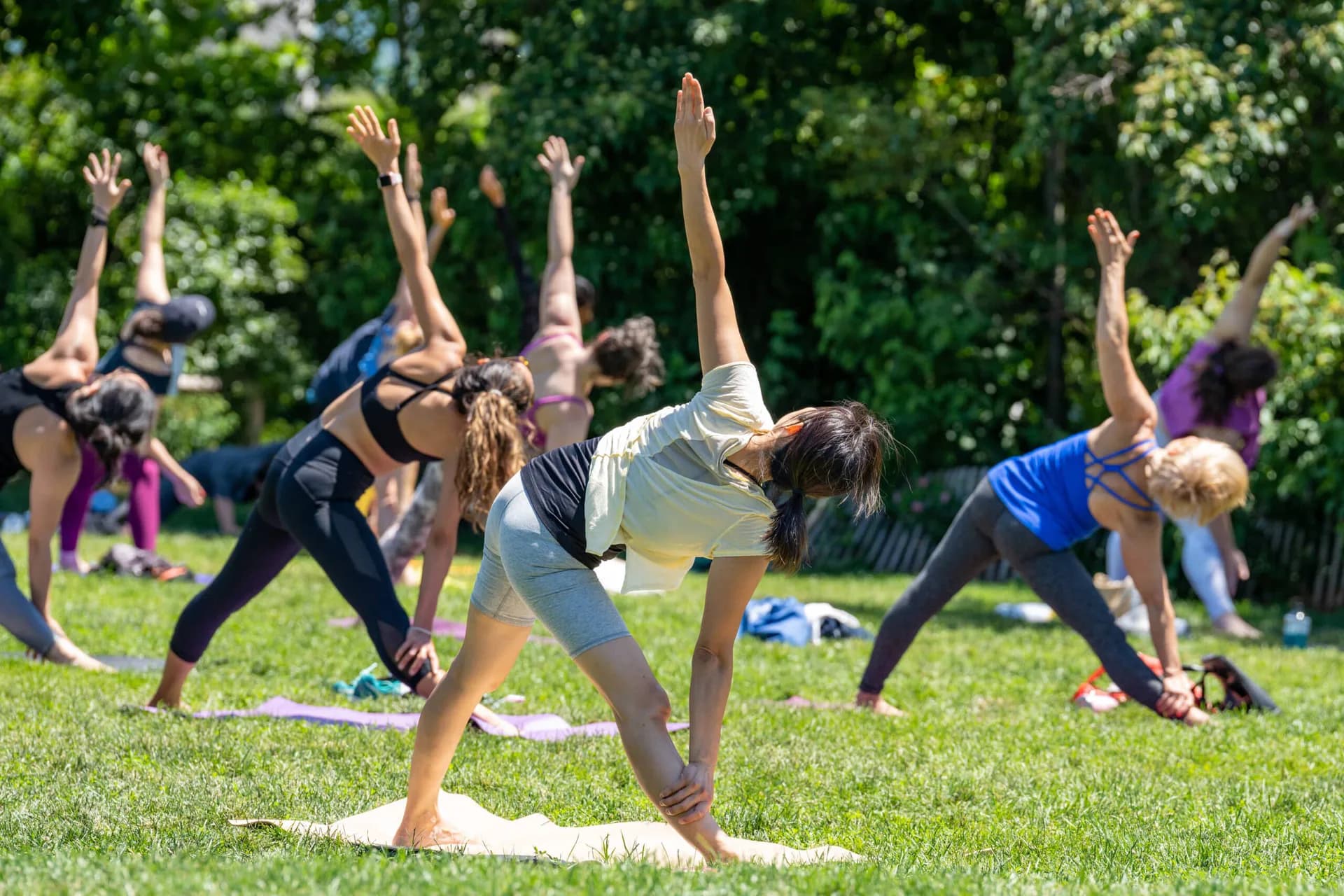 Group of people performing outdoor yoga in a sunny park with lush green foliage, multiple participants in side-stretch poses on mats across grass.