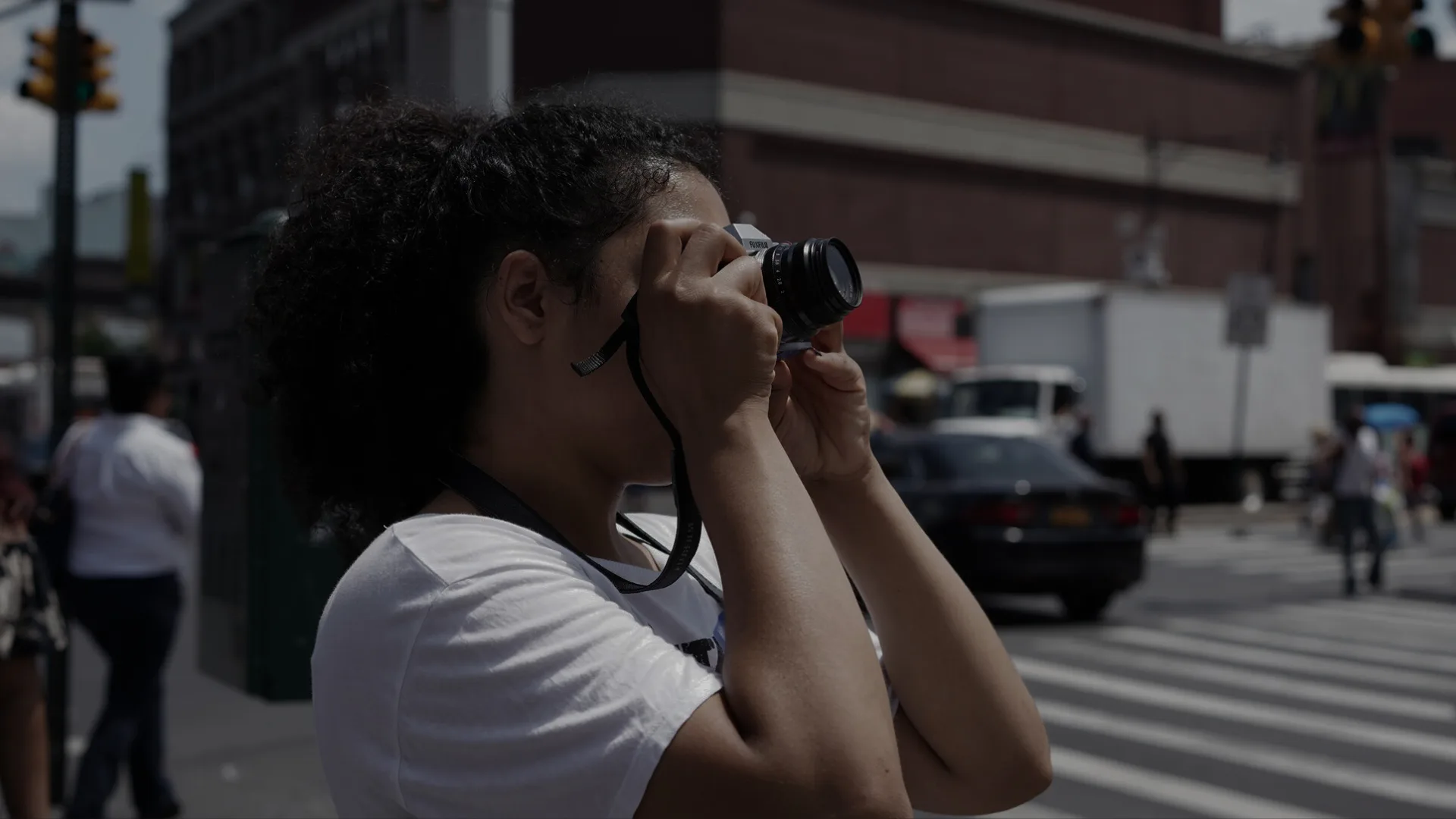 Woman with curly hair holds a camera to her eye photographing a city street with traffic and buildings in the background