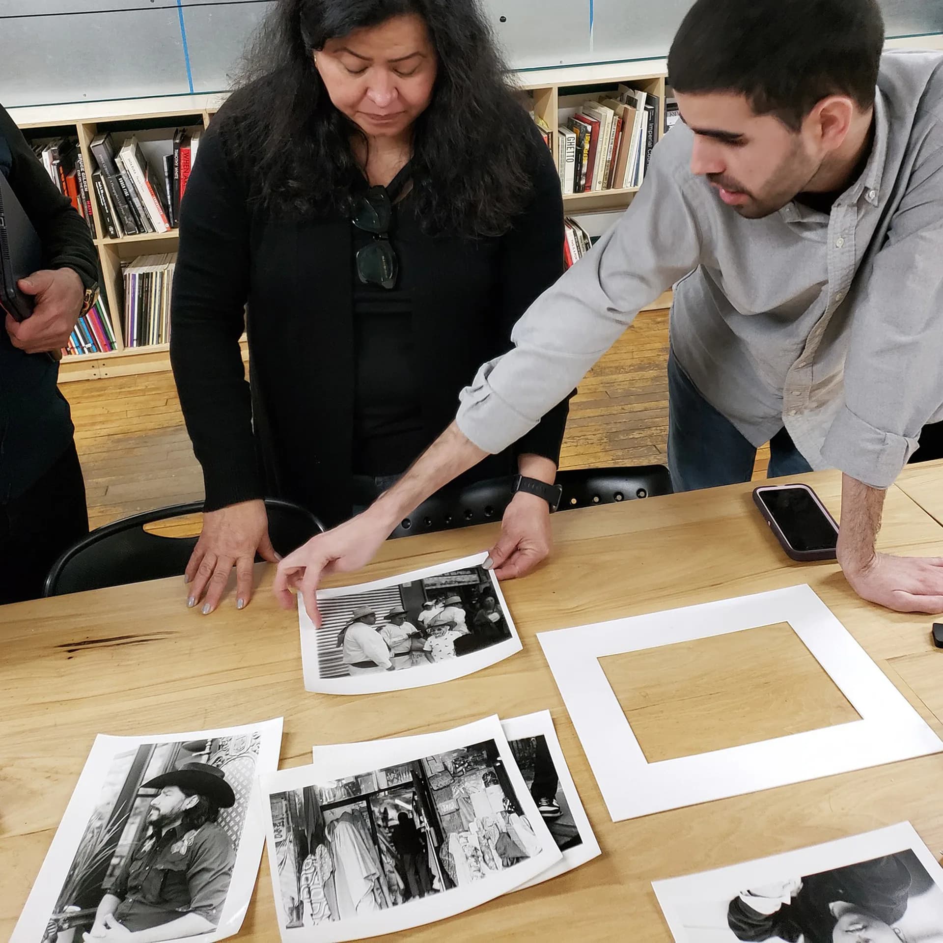 People examine black and white photographic prints laid on a wooden table in a room with bookshelves at the Bronx Documentary Center
