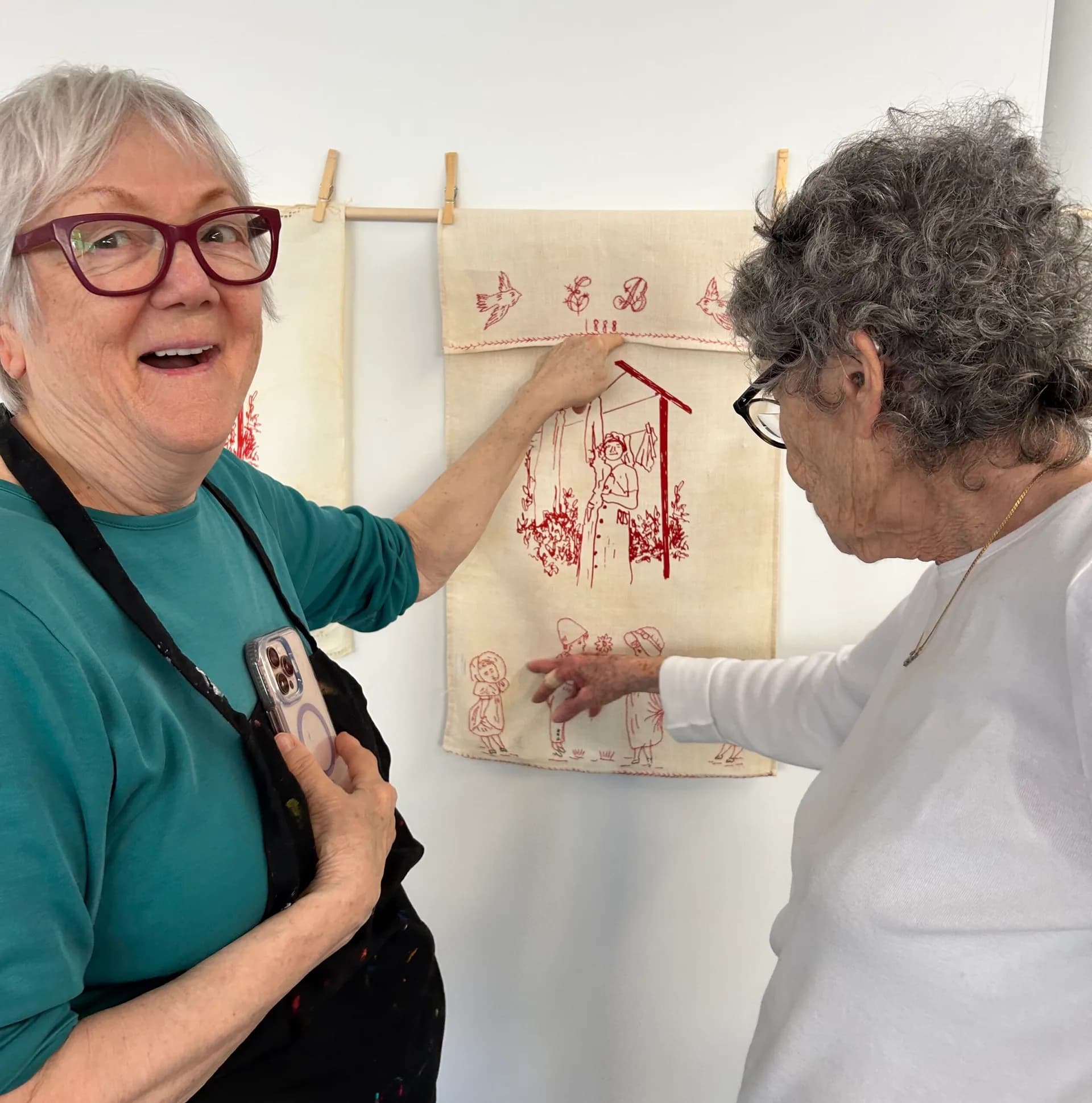 Two people examining a textile artwork hung on a wooden frame; the person on left wears teal and glasses, gesturing toward red embroidered designs on cream fabric.