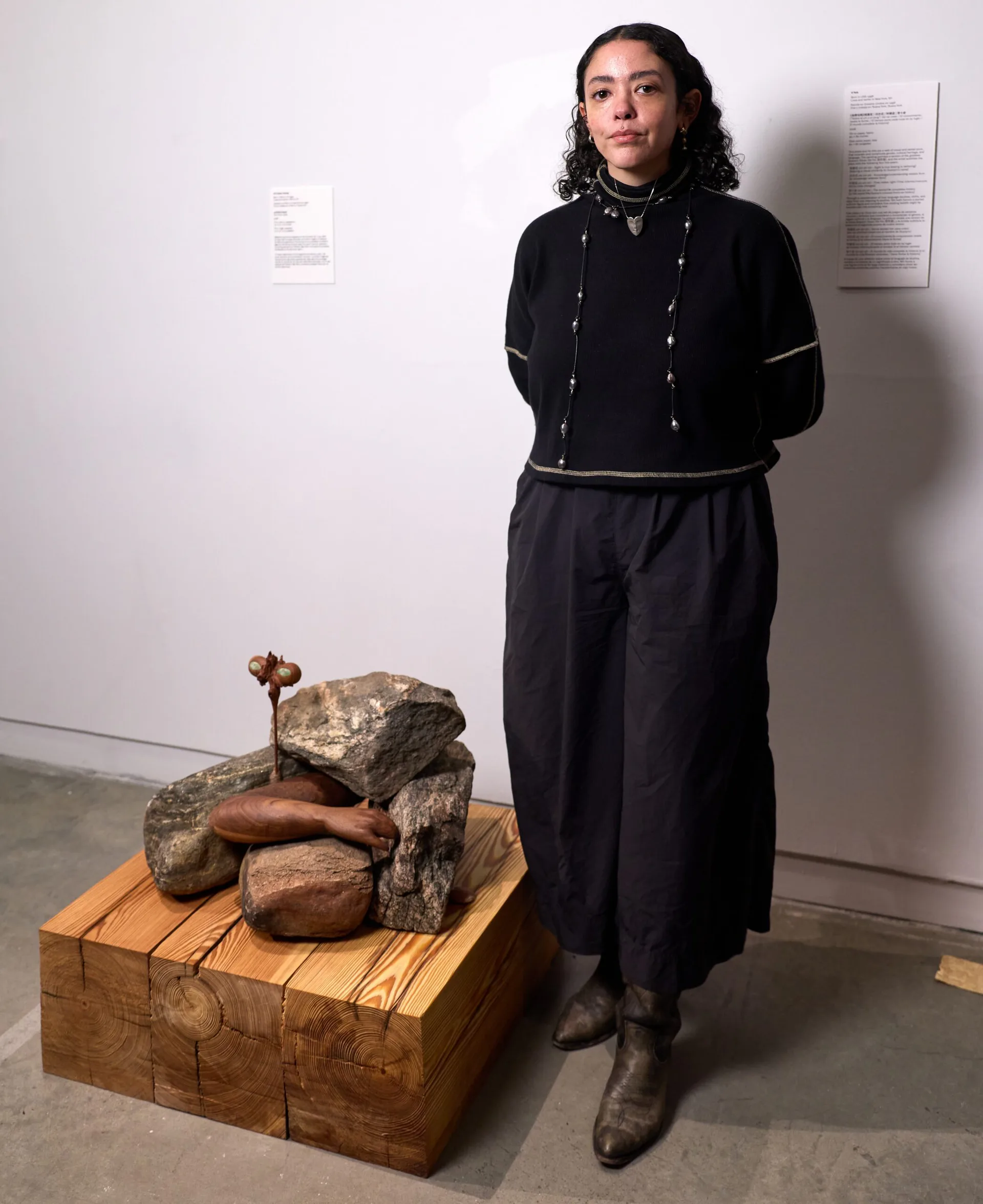 Person in black outfit standing beside a wooden platform displaying stones and a small brass figure in a gallery space with white walls.