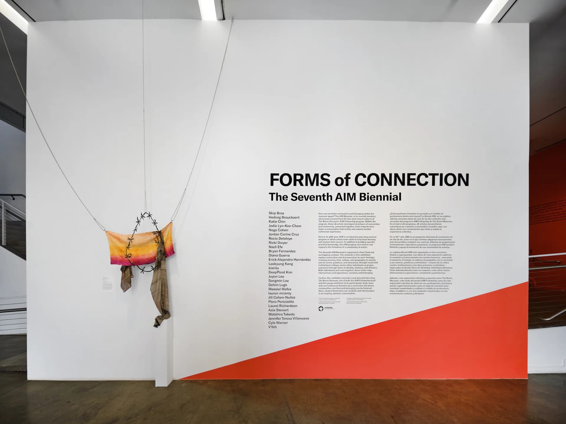 Gallery interior showing a suspended sculptural installation with orange and yellow elements, alongside exhibition signage reading 'FORMS of CONNECTION: The Seventh AIM Biennial' on white and red walls.