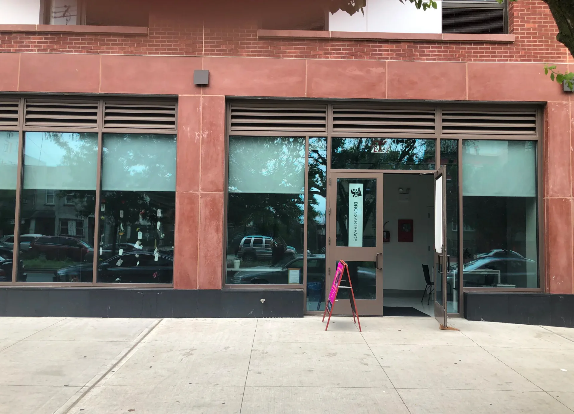 Storefront exterior of BronxArtSpace with brick facade, glass windows, and open entrance door with a pink sandwich board sign on the sidewalk