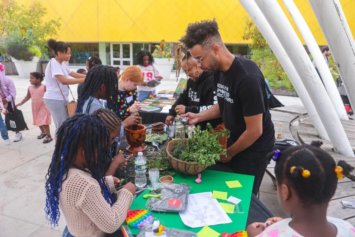 Man facilitating a colorful craft activity at Brooklyn Children's Museum with children at a green table under yellow canopy outdoors.