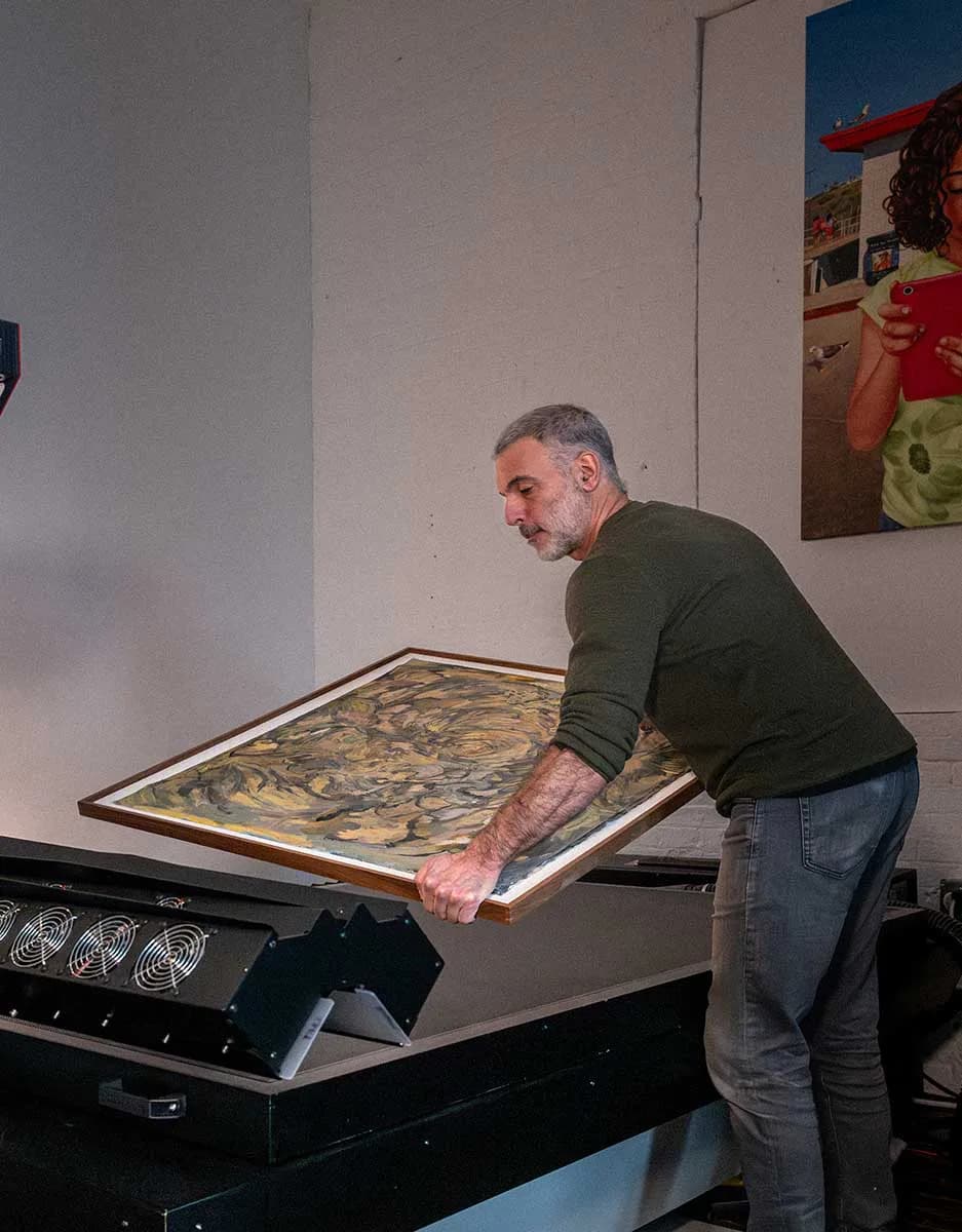Man lifting or tilting a large framed artwork with layered gold and brown tones in a gallery or workshop setting.