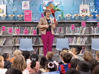 Library staff member in colorful outfit leading storytelling session with children seated on floor in front of shelves of books.
