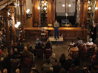 Person speaking on stage in ornate historic hall with wooden details and chandeliers, audience seated facing the speaker.
