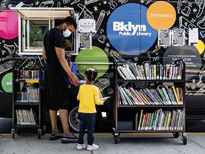 Adult and child browsing books at mobile Brooklyn Public Library kiosk with blue circular signage and black patterned backdrop.