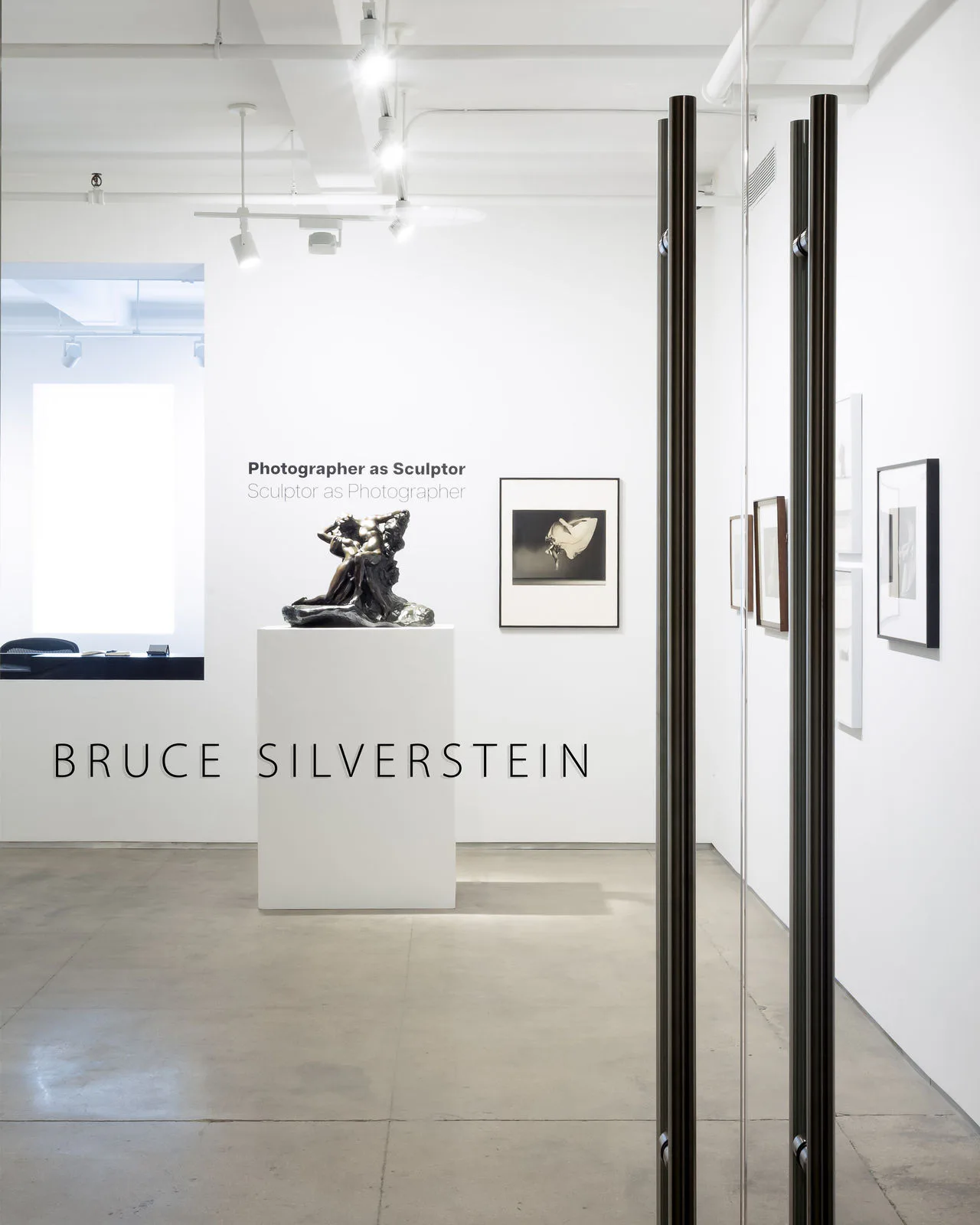 Interior of Bruce Silverstein gallery with white walls, a sculpture on a pedestal, framed photographs, and glass doors with black handles in the foreground