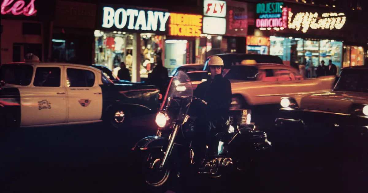 Nighttime color photograph of a motorcycle parked on an urban street with neon storefront signage including 'BOTANY' visible in the background.