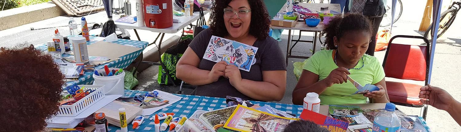 Adults and children at outdoor table with art supplies and colorful printed materials at BxArts Factory event or workshop.