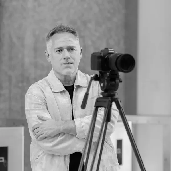 Black and white portrait of a man in light-colored shirt posing with a camera on tripod.