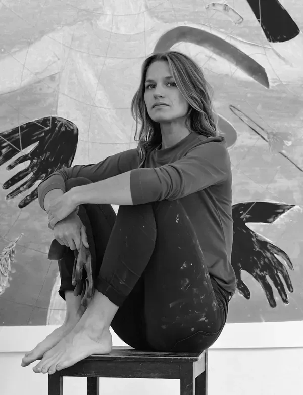 Black and white portrait of a woman sitting on bench, interacting with painted hands and wings on wall behind her.
