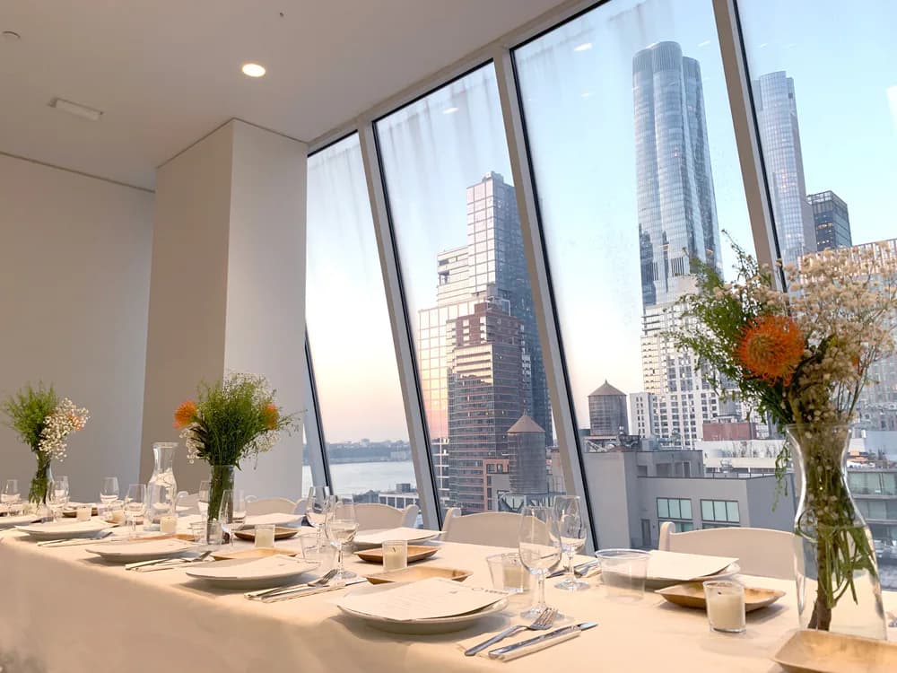 Bright, minimalist dining room with floor-to-ceiling windows overlooking a city skyline with tall buildings and water; white table settings and fresh flowers in glass vases.