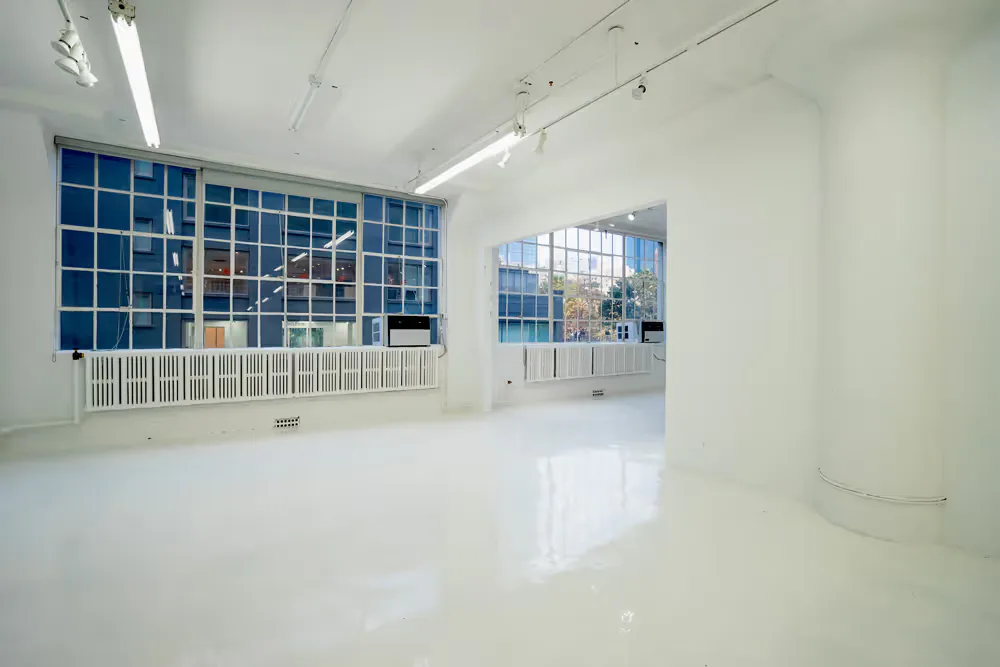 Clean, empty white gallery interior with large blue-tiled windows reflecting natural light; polished concrete floor and minimal architectural features.