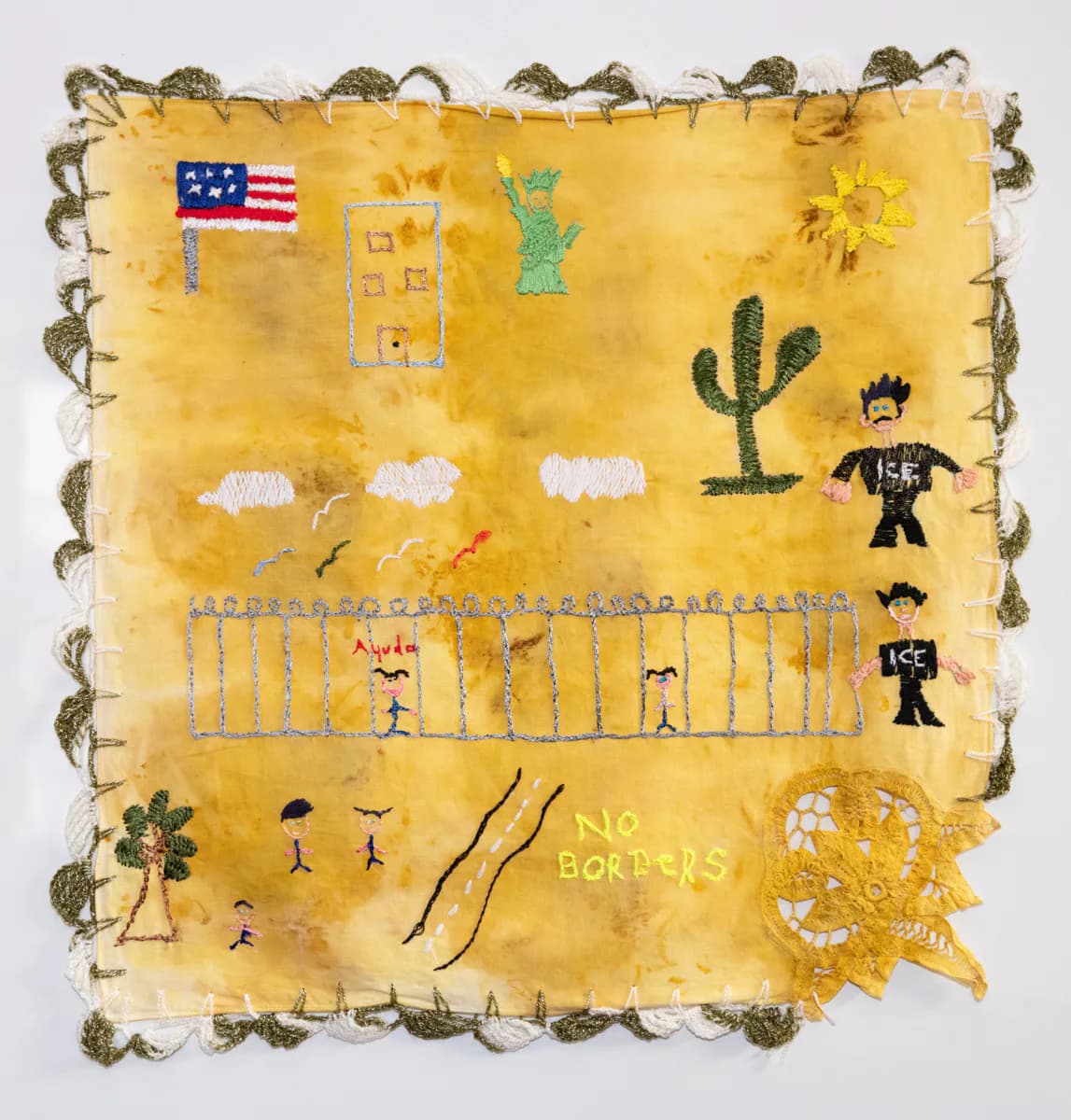 Mixed-media textile artwork on mustard-yellow fabric with raw edges and decorative border; features childlike drawings of American flag, cactus, stick figures, white clouds, and text "NO BORDERS" in naïve style.
