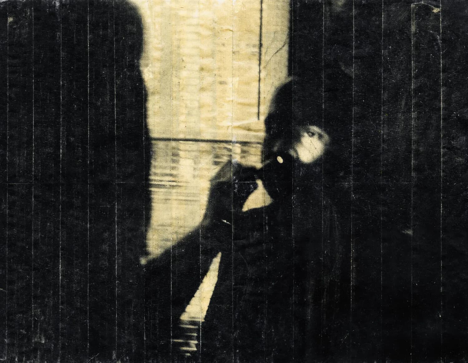 Black and white photograph with heavy film grain showing figure in shadows and geometric light patterns on dark textured wall; high-contrast, moody composition.