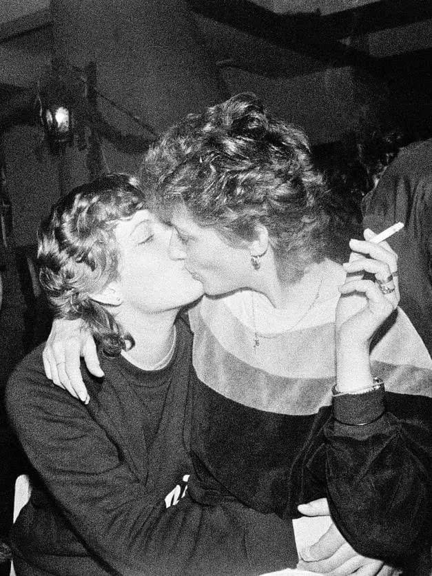 Black and white photograph of two people kissing in profile, both holding cigarettes, captured in an intimate moment indoors.