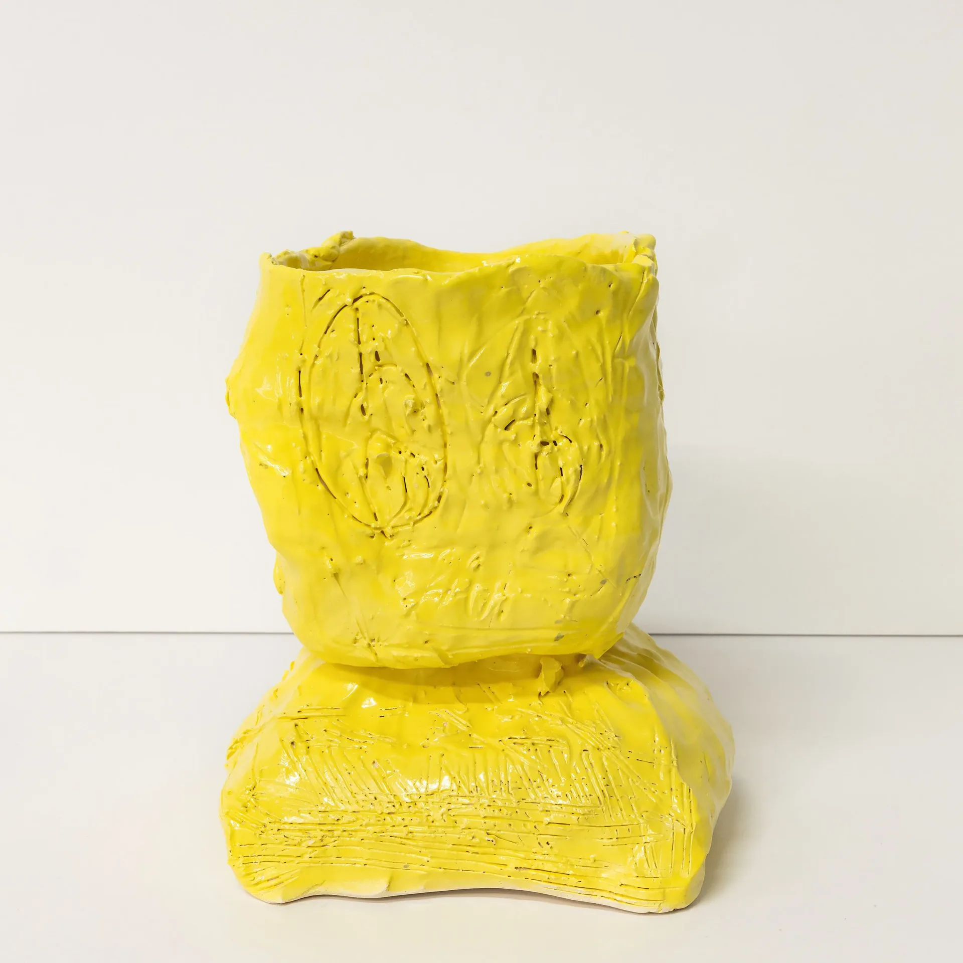 Bright yellow sculptural form with textured surface and carved eye motifs, stacked pedestal structure.