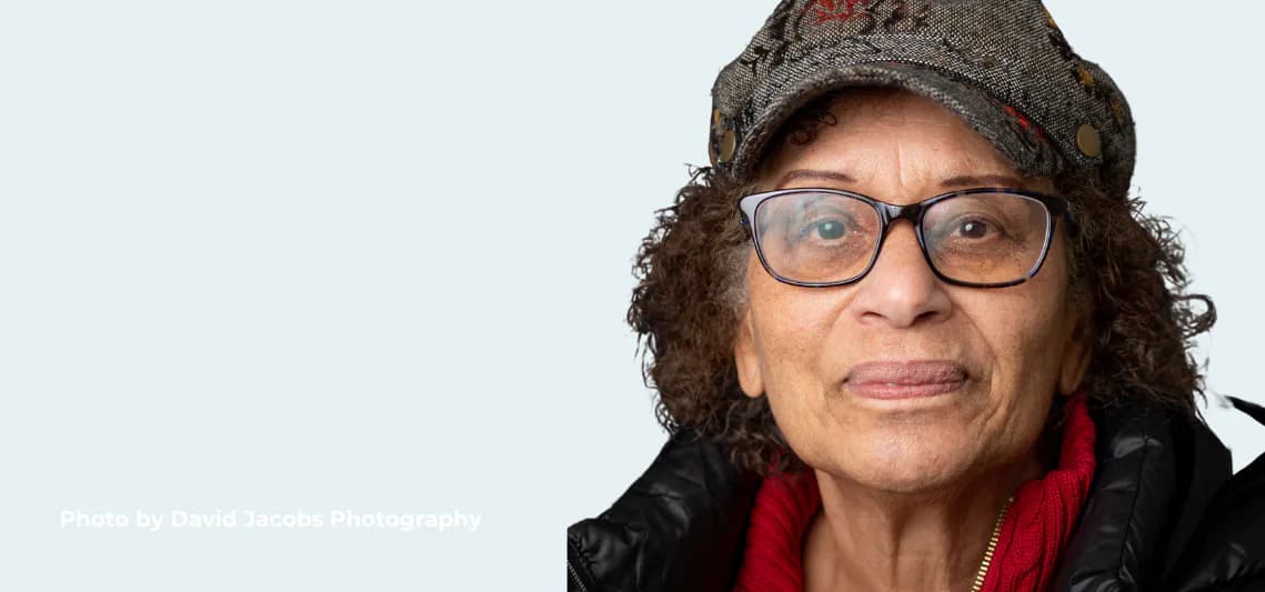 Person wearing glasses, a gray knit cap, and black jacket with red scarf, smiling at camera against light blue background.