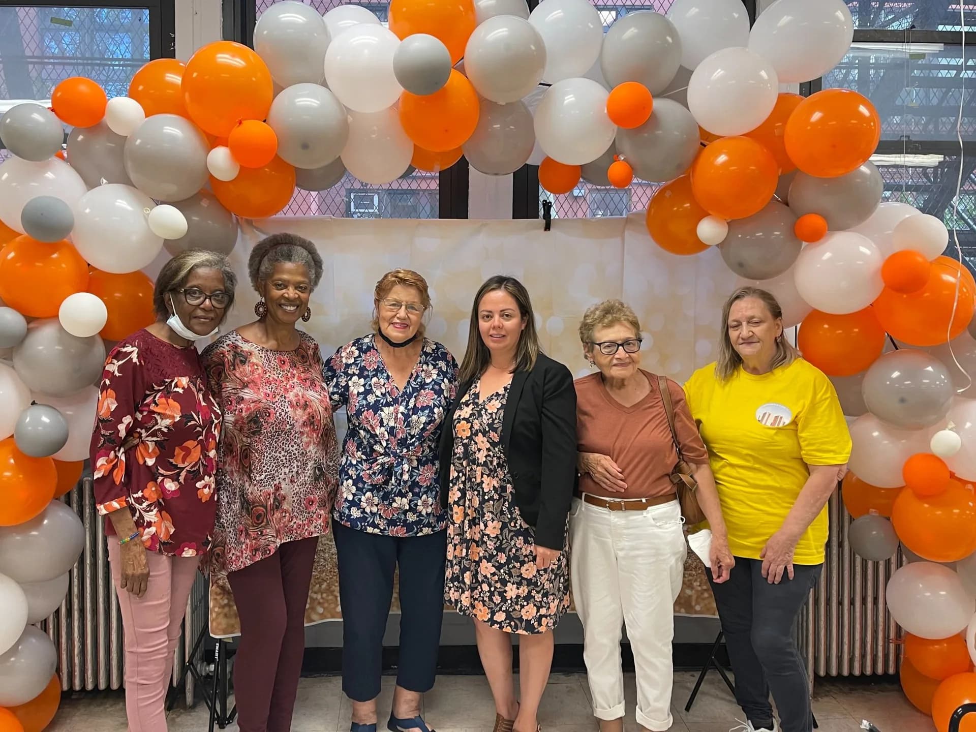 Six people standing in front of orange, white, and gray balloon arch decoration in an indoor event space or gallery.