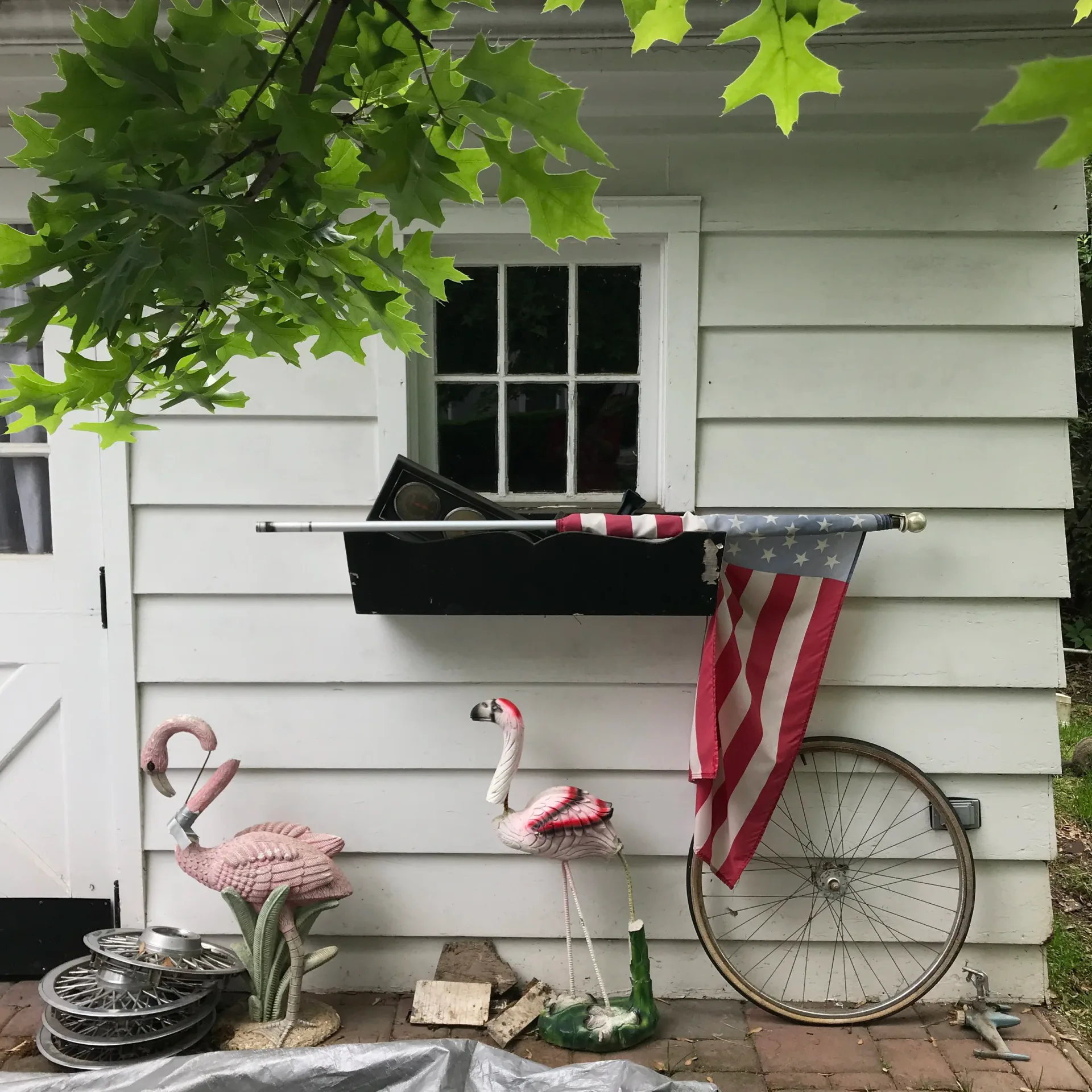 White wooden exterior wall with black window, decorated with pink flamingo sculptures, an American flag, bicycle wheel, and potted plants.