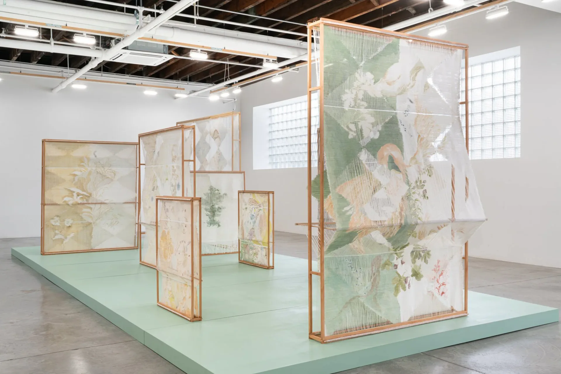Mixed-media installation with copper-framed translucent panels displaying botanical and figurative imagery, arranged on a mint-green platform in a gallery.