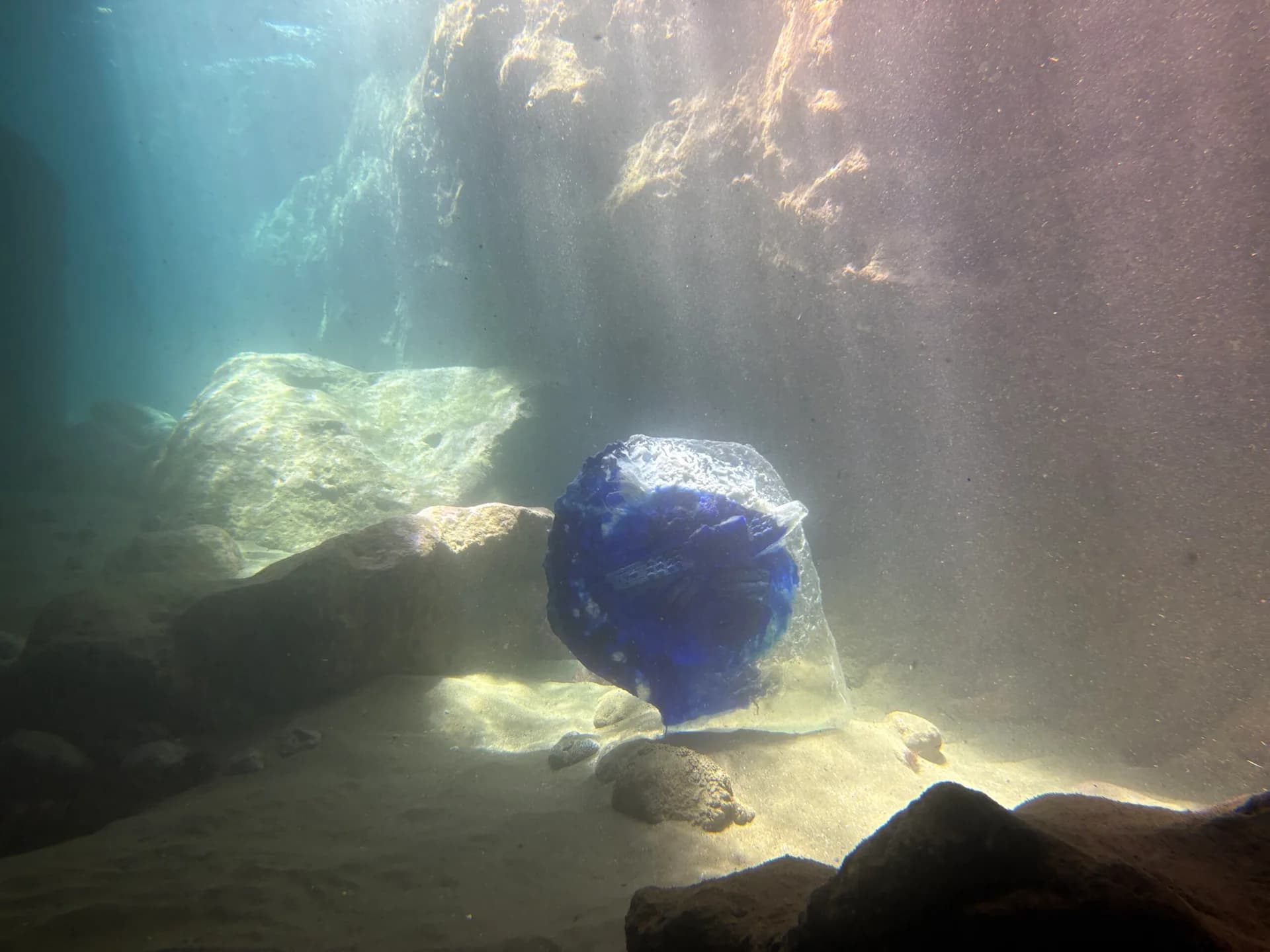 Underwater photograph with blue spherical object resting on rocky bottom, filtered sunlight illuminating the scene.