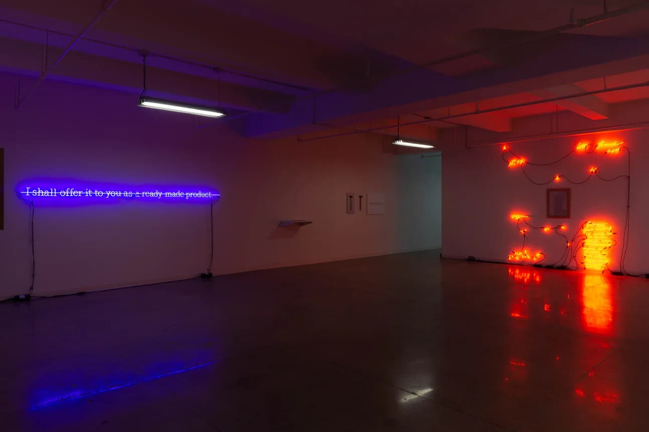 Gallery interior with blue neon text on left wall, red and orange neon lights on right side, polished concrete floor, fluorescent ceiling fixtures.