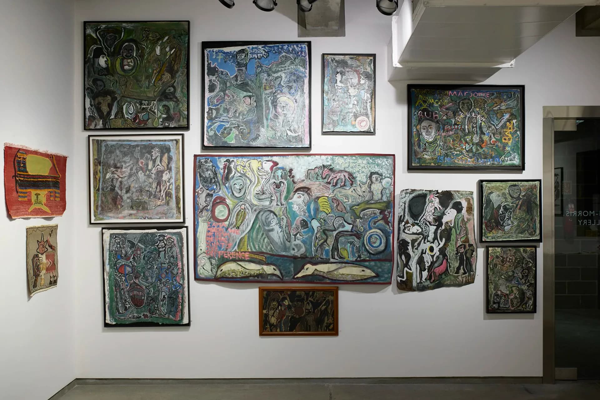 Cavin-Morris Gallery wall display featuring numerous framed artworks in varied styles, with densely arranged paintings and drawings covering the white wall.