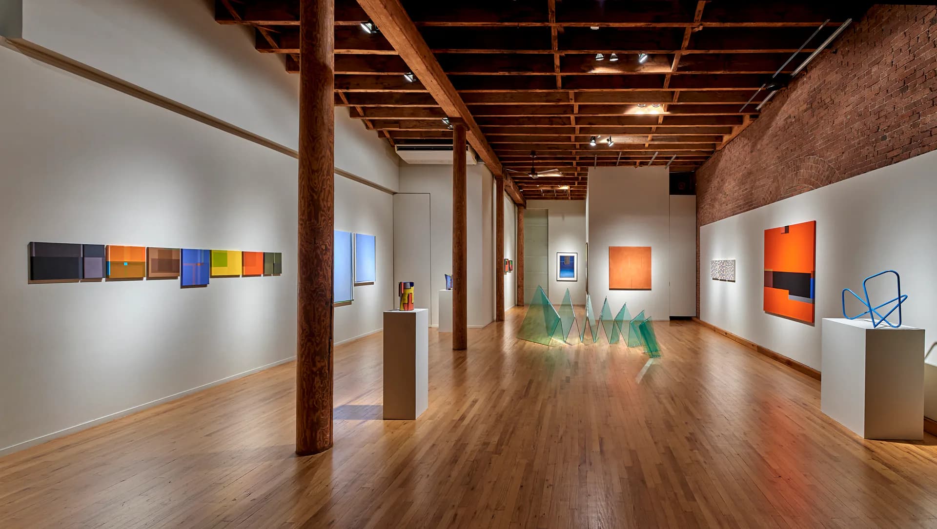 Gallery interior with wooden floors, exposed beams, and white walls displaying colorful abstract artworks and sculptures on pedestals.