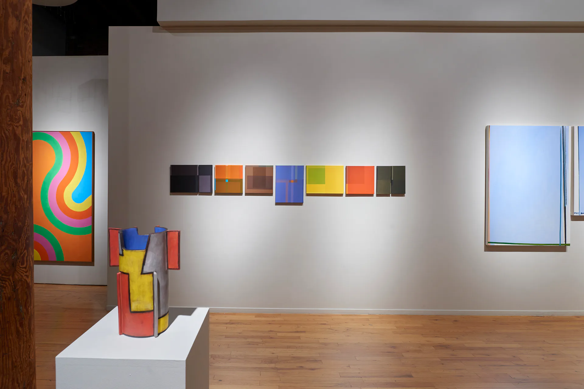 Gallery corner showing abstract paintings in bright colors and a sculptural form on a white pedestal against white walls.