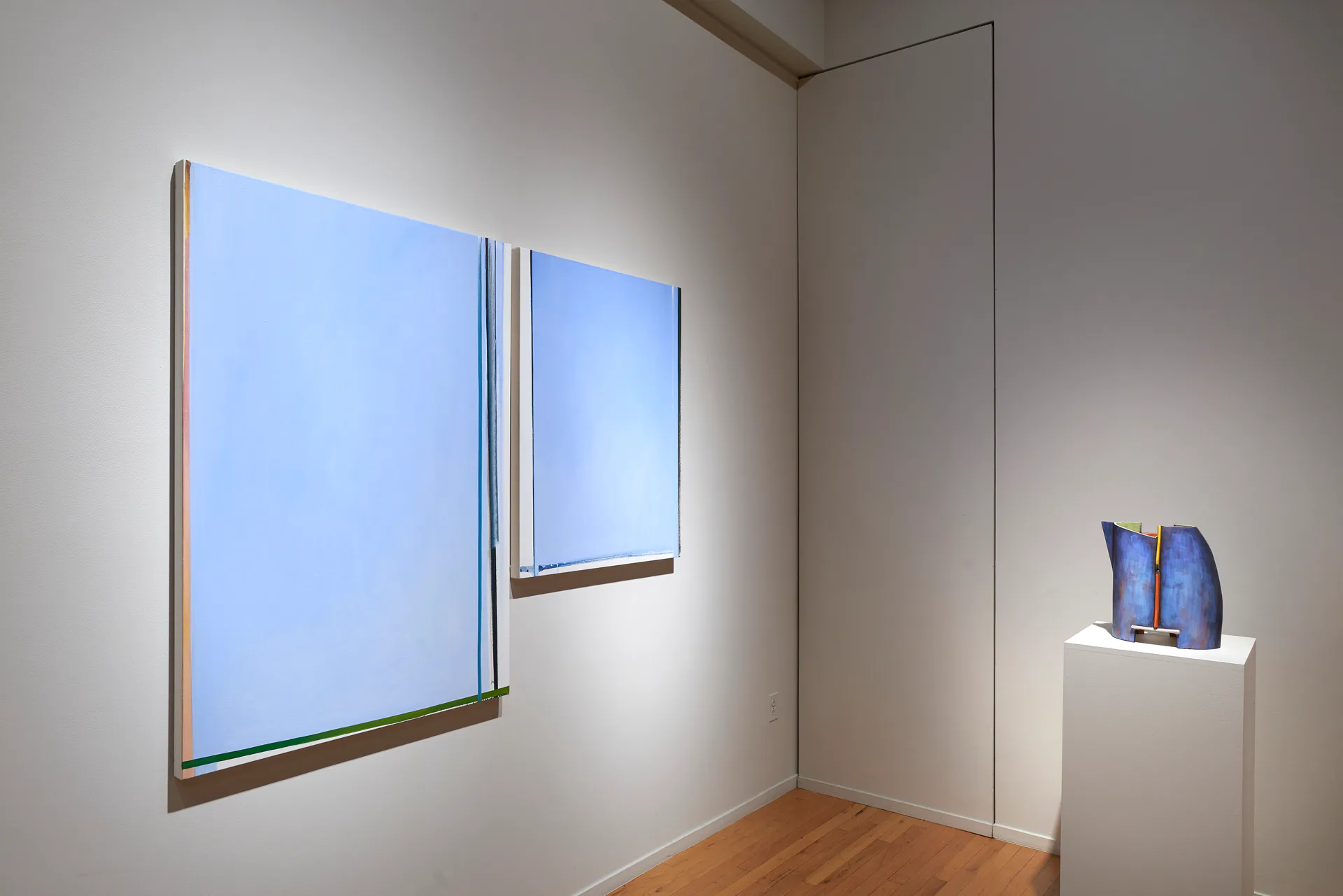 Corner gallery space with wooden floor, white walls, two large light-blue framed artworks, and a sculptural piece on a pedestal.