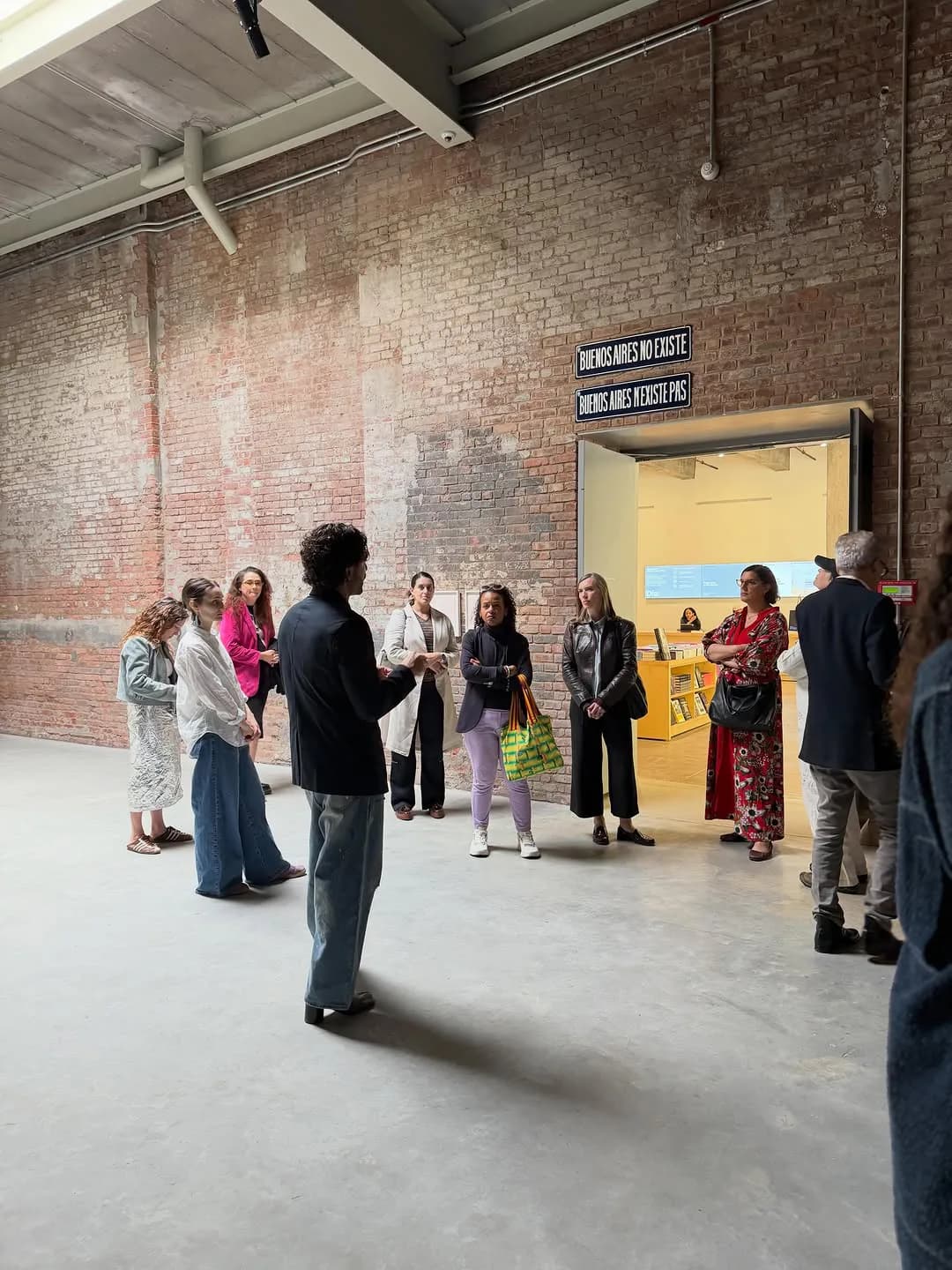 Visitors chat casually in a large industrial gallery space with exposed brick walls and concrete floor, near a sign reading 'Buenos Aires no existe'.