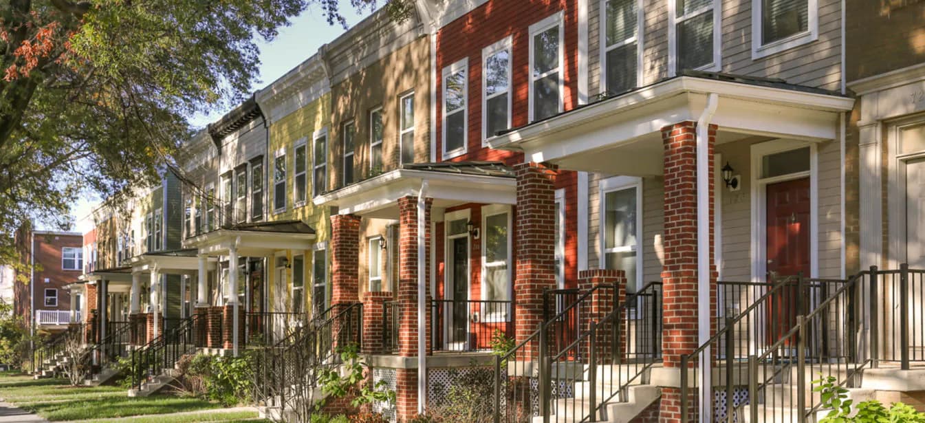 Tree-lined street of historic rowhouses with varied brick and painted facades, wrought-iron railings, and storefronts.