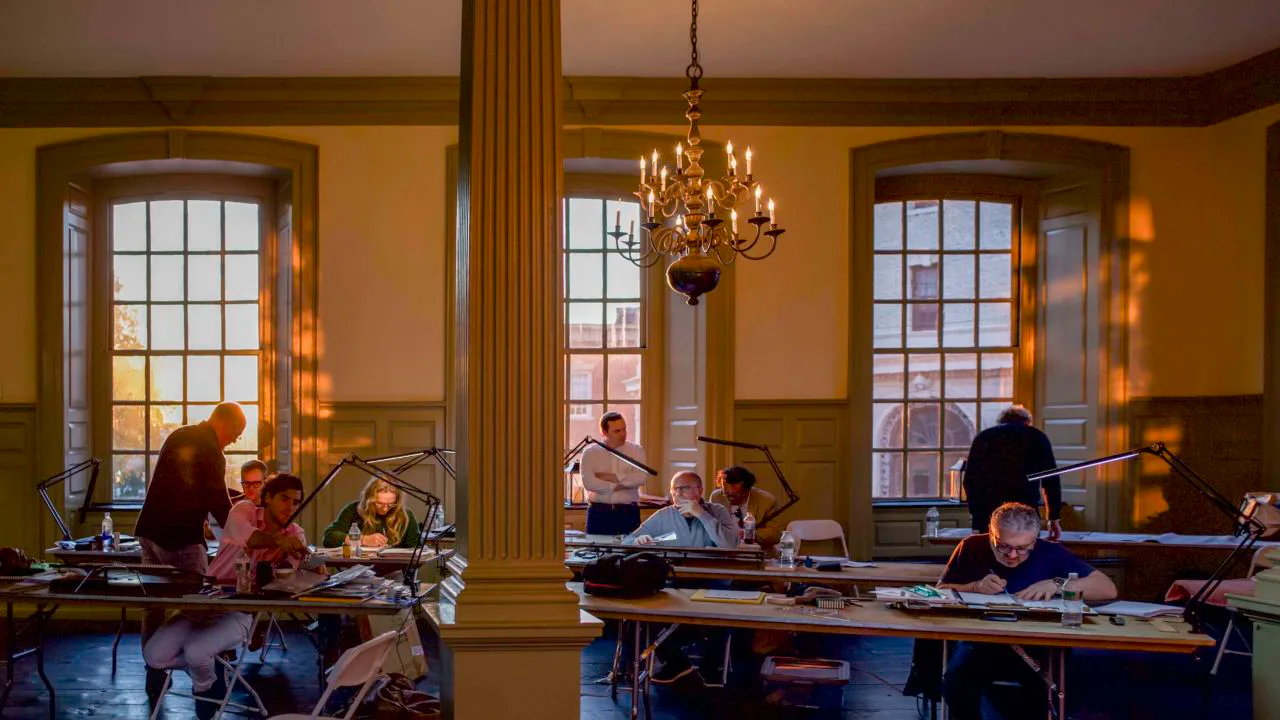 Classical interior with yellow walls, fluted columns, chandelier, tall windows, and people working at tables in warm light.