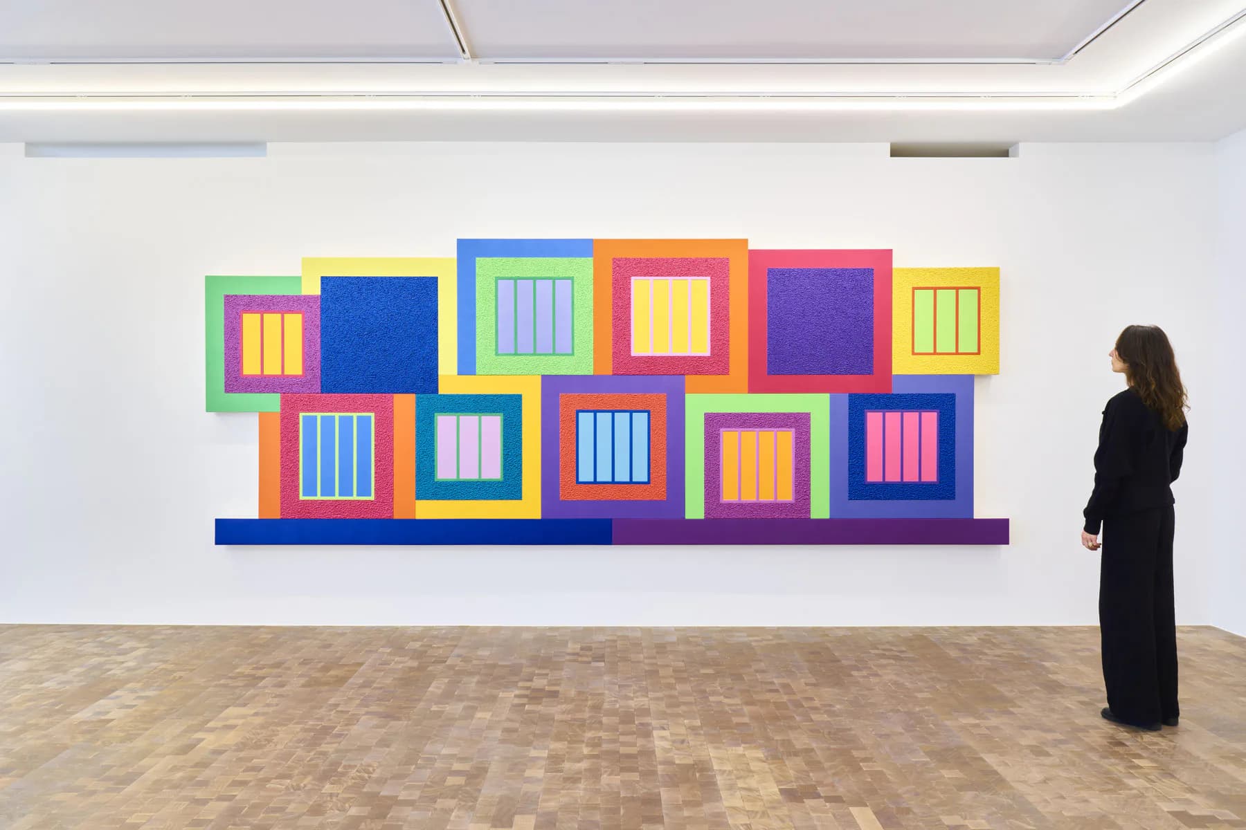 Gallery interior with white walls and wooden floor; a person in black views a large colorful mural of stacked geometric squares in bright primary colors.