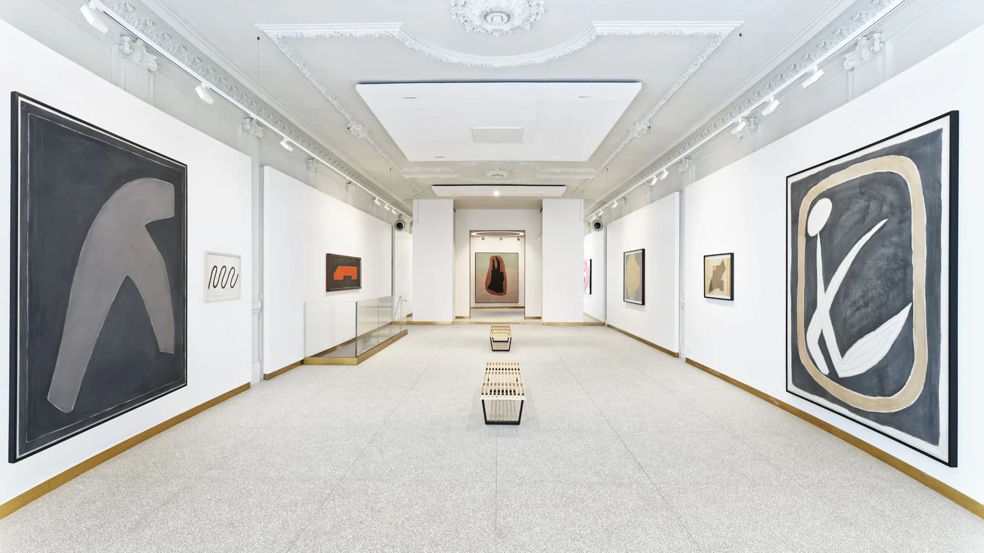 Gallery corridor with bright white walls, spotlit artworks displayed on both sides, and clean concrete floor extending toward distant doorway.