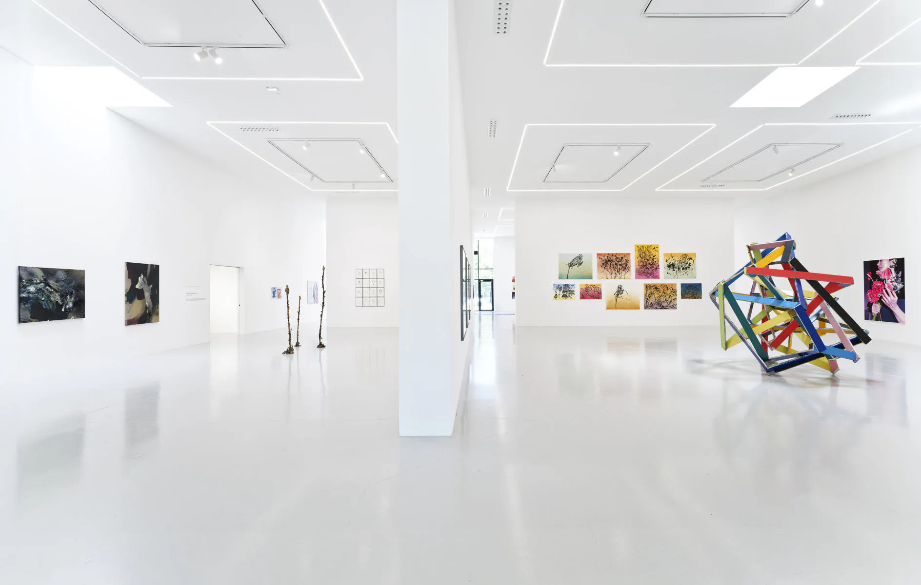 Modern white gallery interior with minimalist artwork displayed on walls and a colorful sculptural installation in center foreground.