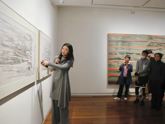 Gallery interior with woman gesturing toward large graphite drawing on left wall; visitors viewing colorful artwork on right.
