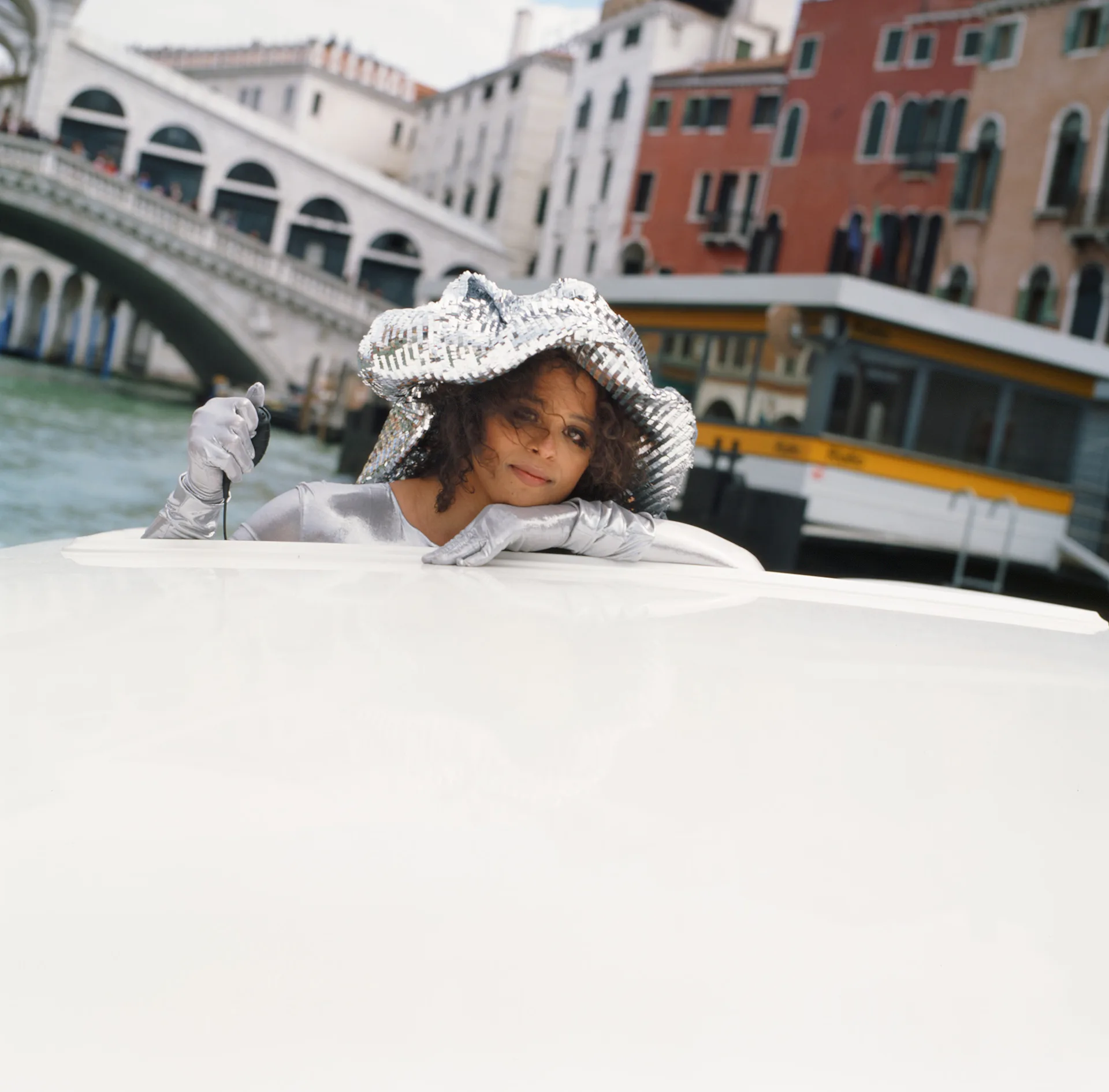Child in white hooded coat peers over white surface with Venice canal, Rialto Bridge, and colorful buildings in soft-focus background.