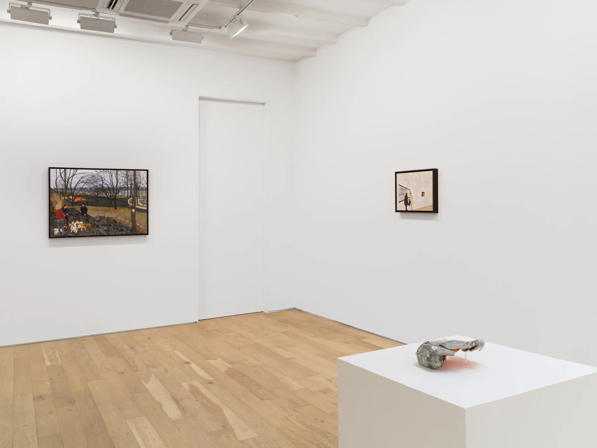Minimal gallery space with two small framed artworks on walls and sculptural object on white pedestal.