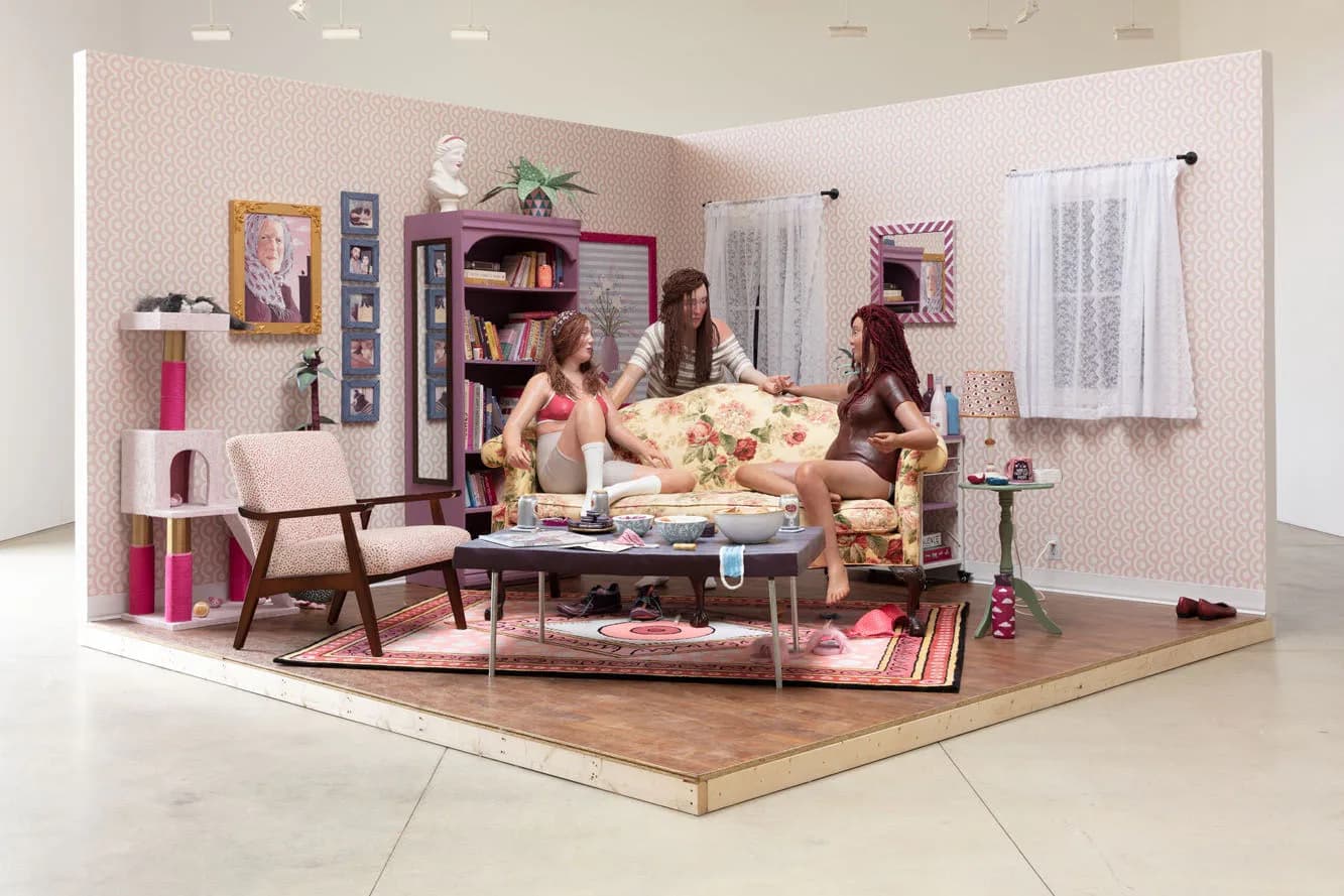 Diorama-style installation depicting a miniature living room with figurines, furniture, and pink wallpaper in a white gallery setting.