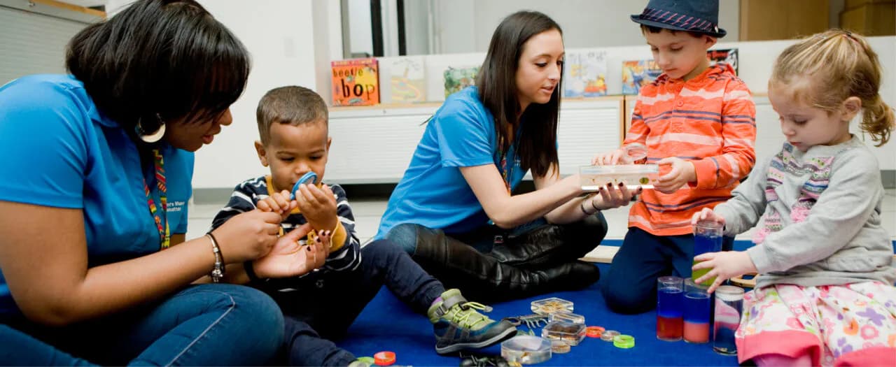 Two museum educators in blue shirts guide children in hands-on play with animal figures and colorful blocks on a blue mat.