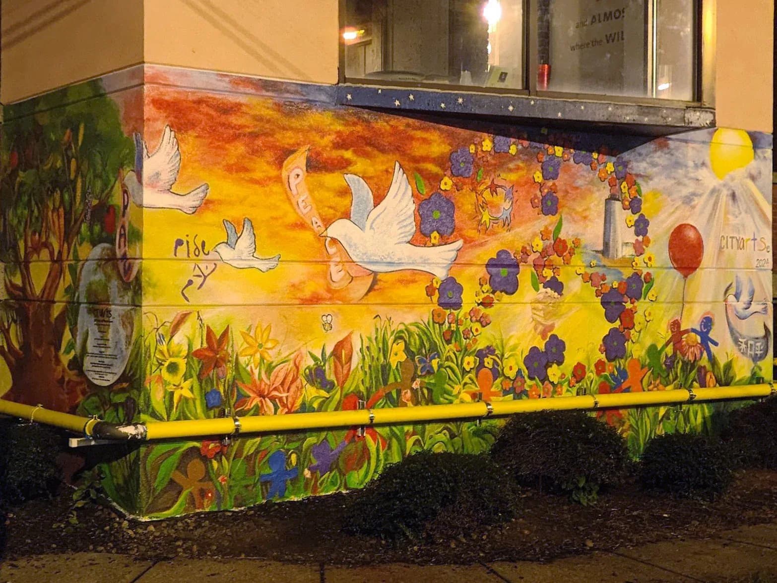 Colorful mural on a utility box featuring white doves, orange sun, yellow flowers, green grass, and diverse figures in warm and cool tones.