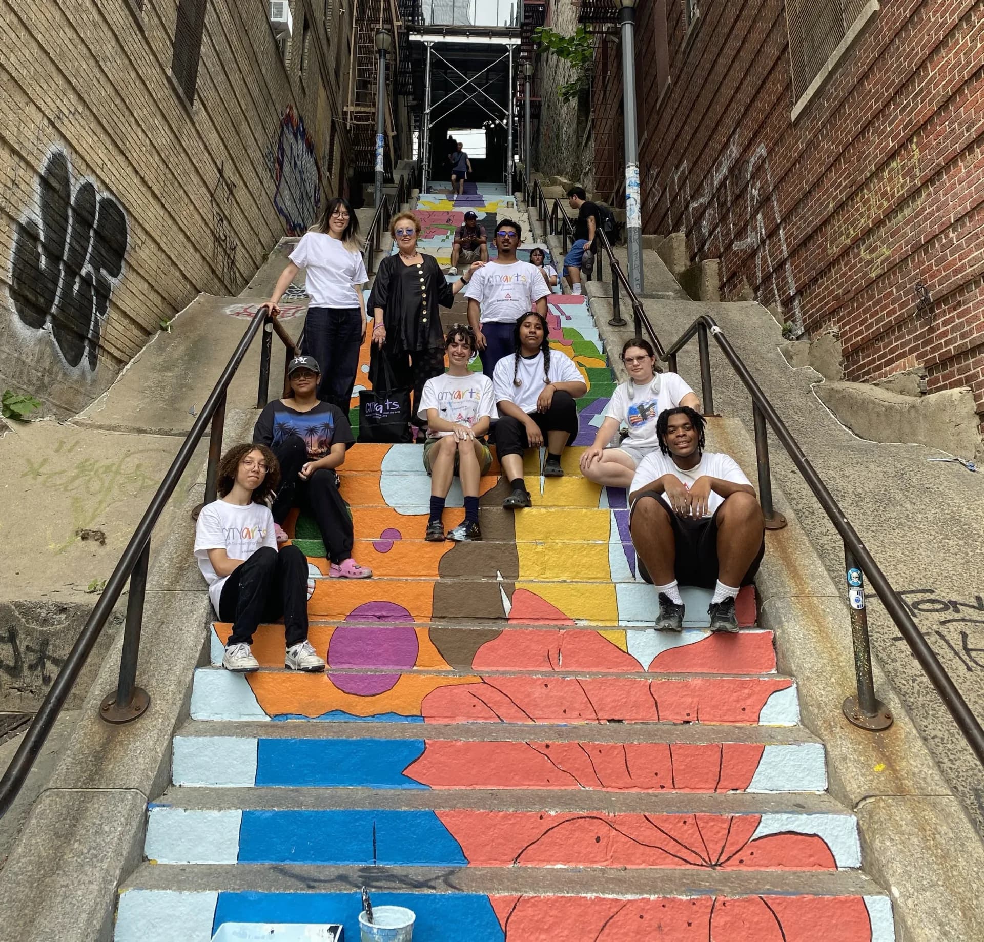 Group of young people posing on an outdoor staircase painted with colorful geometric shapes in orange, blue, red, and yellow.