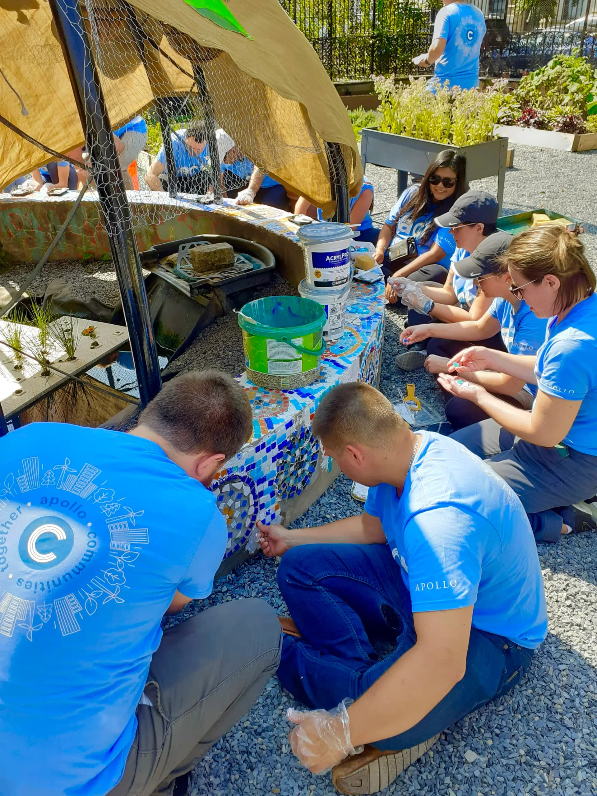 People in blue CITYarts shirts engaged in a hands-on activity under a tent with paint materials and water containers visible.