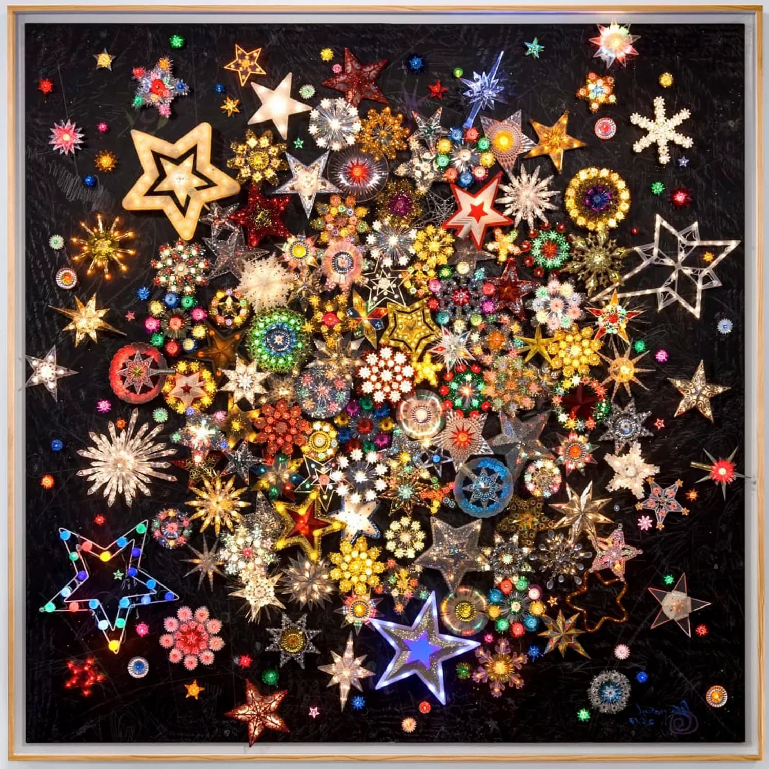 Assemblage artwork on black background composed of hundreds of colorful decorative beads, rhinestones, and star ornaments arranged in a dense, radiating composition with wooden frame.