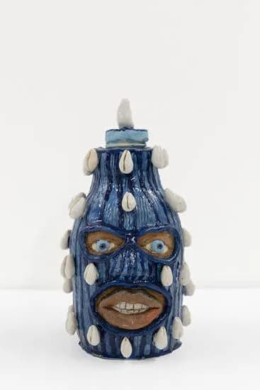Blue ceramic vessel with white circular studs, topped with a pointed finial, featuring a stylized face with large eyes and prominent grin.